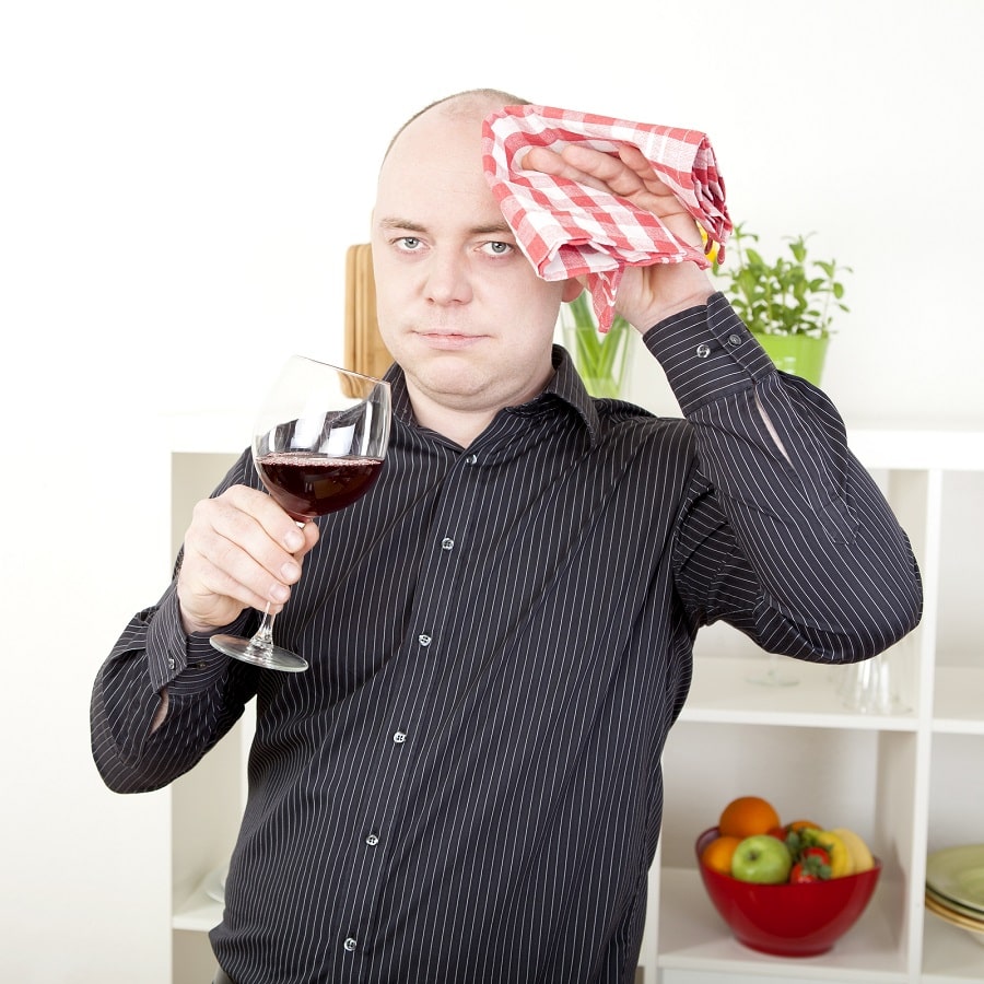 Mann hält sich Kopf mit Tuch und einem Rotweinglas um Schmerzempfindlichkeit durch Alkohol vor Haartransplantation zu demonstrieren