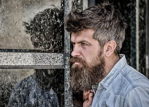 Eine Barthaartransplantation hilft bei Haarschwund im Gesicht Mann mit Vollbart und Hemd posiert seitlich angelehnt an einer Wand