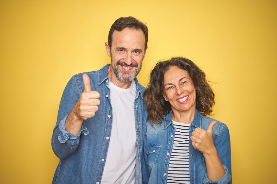 Ein Mann und eine Frau stehen nebeneinander vor einem gelben Hintergrund und zeigen ihren daumen